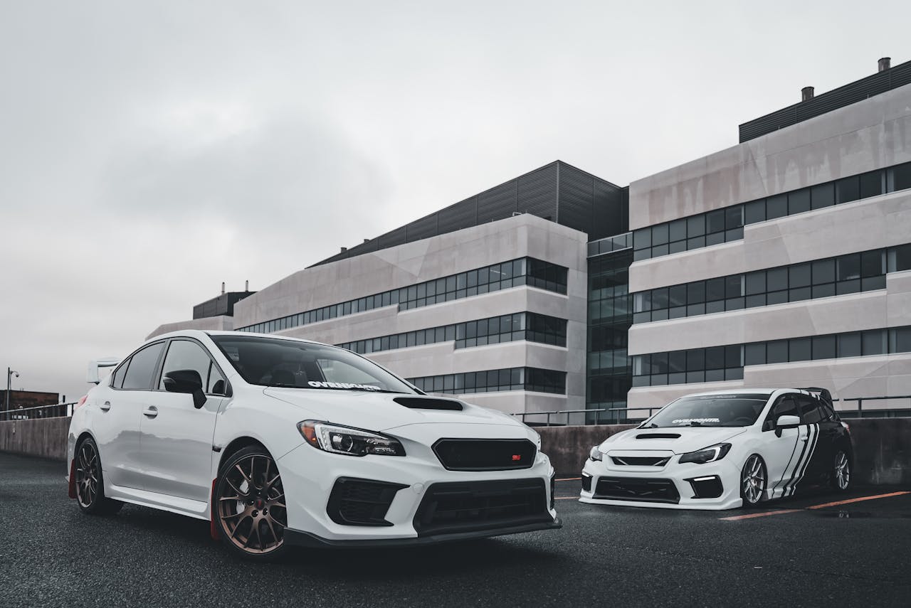 Two tuned white Subaru WRX sports cars parked in a modern urban environment with building backdrop.